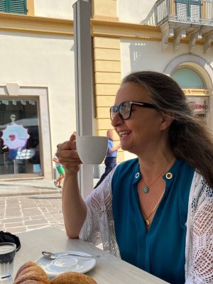 Auf dem Foto sieht man Manuela Paolini. Sie sitzt in einer italienischen Bar zum Frühstück mit einem Cappuccino und Cornetto draußen an einem Tisch und genießt das Leben.
