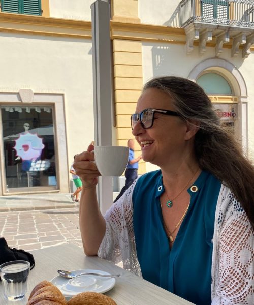 Auf dem Foto sieht man Manuela Paolini. Sie sitzt in einer italienischen Bar zum Frühstück mit einem Cappuccino und Cornetto draußen an einem Tisch und genießt das Leben.